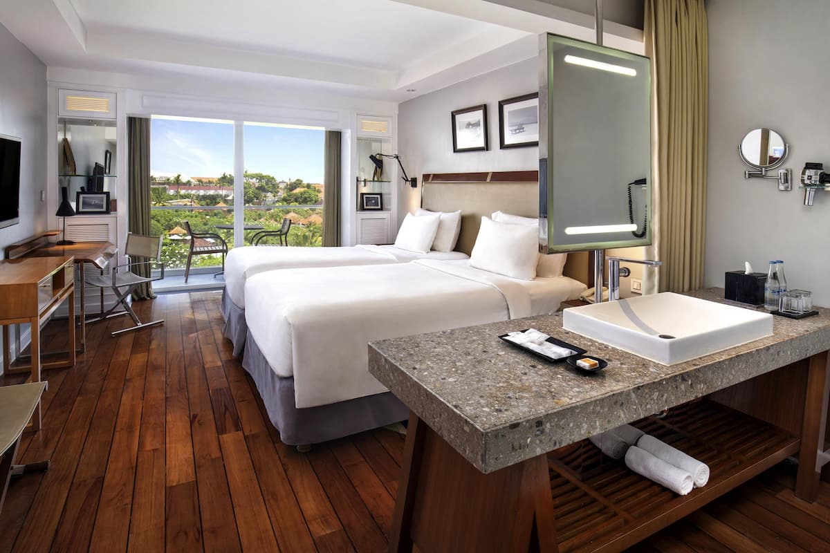 A hotel room with two beds, a desk, a TV, a bathroom with a sink, and a view of greenery.