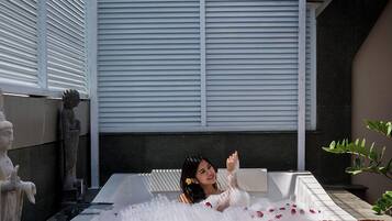 A person relaxing in a large, white hot tub with rose petals scattered around.