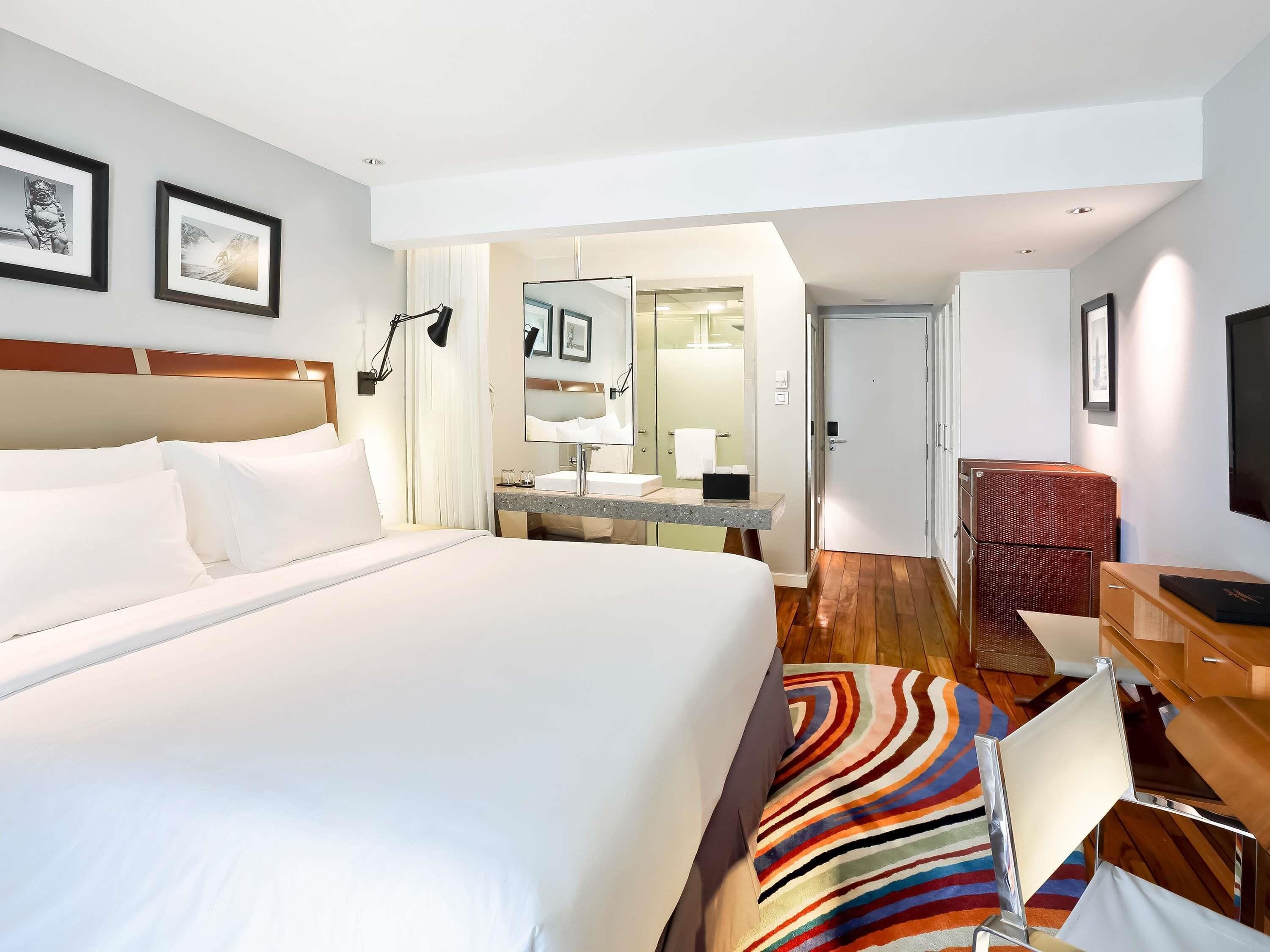 A hotel room with a large bed, a desk with a mirror, a television, and a colorful rug.