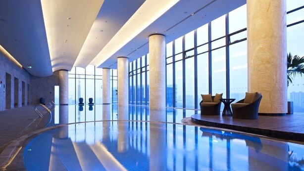Una piscina interior moderna con paredes de vidrio transparente, un área de descanso con sillones y vistas al horizonte de la ciudad.