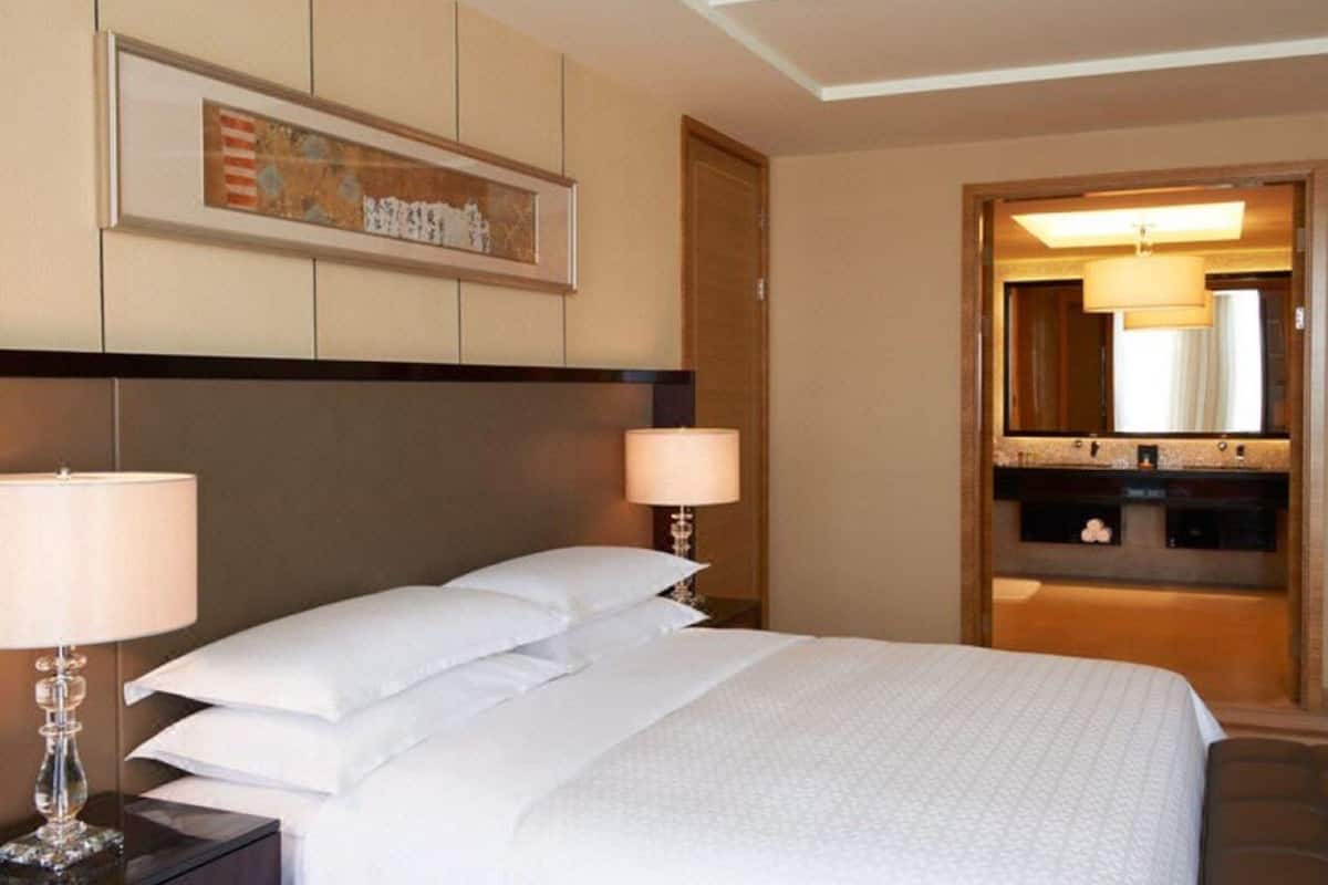 A hotel room with a large bed, two bedside lamps, a framed artwork above the bed, and a bathroom visible through an open door.