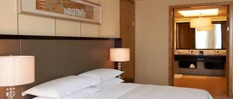 A hotel room with a large bed, two bedside lamps, a framed artwork above the bed, and a bathroom visible through an open door.