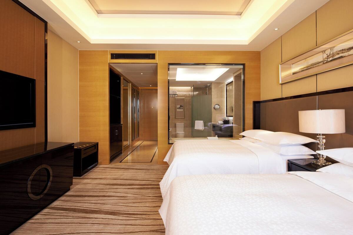A hotel room with two beds, a TV, a mirror, and a bathroom visible through an open door.