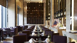 Un restaurante moderno con mesas de madera oscura y sillas moradas, una barra larga con exhibición de alimentos y una pared con diseño en el fondo.