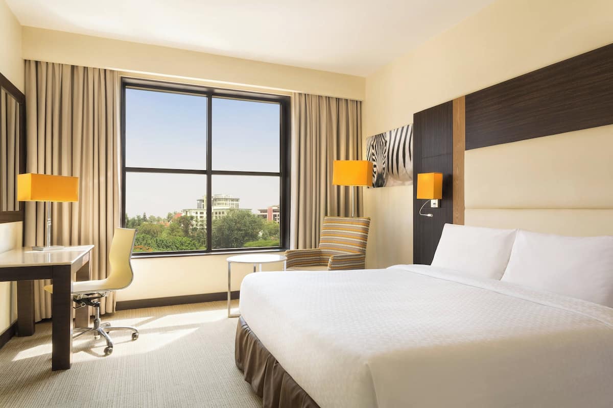 Ein Hotelzimmer mit einem großen Bett, einem Schreibtisch, einem Stuhl und Blick auf die Stadt durch ein großes Fenster.