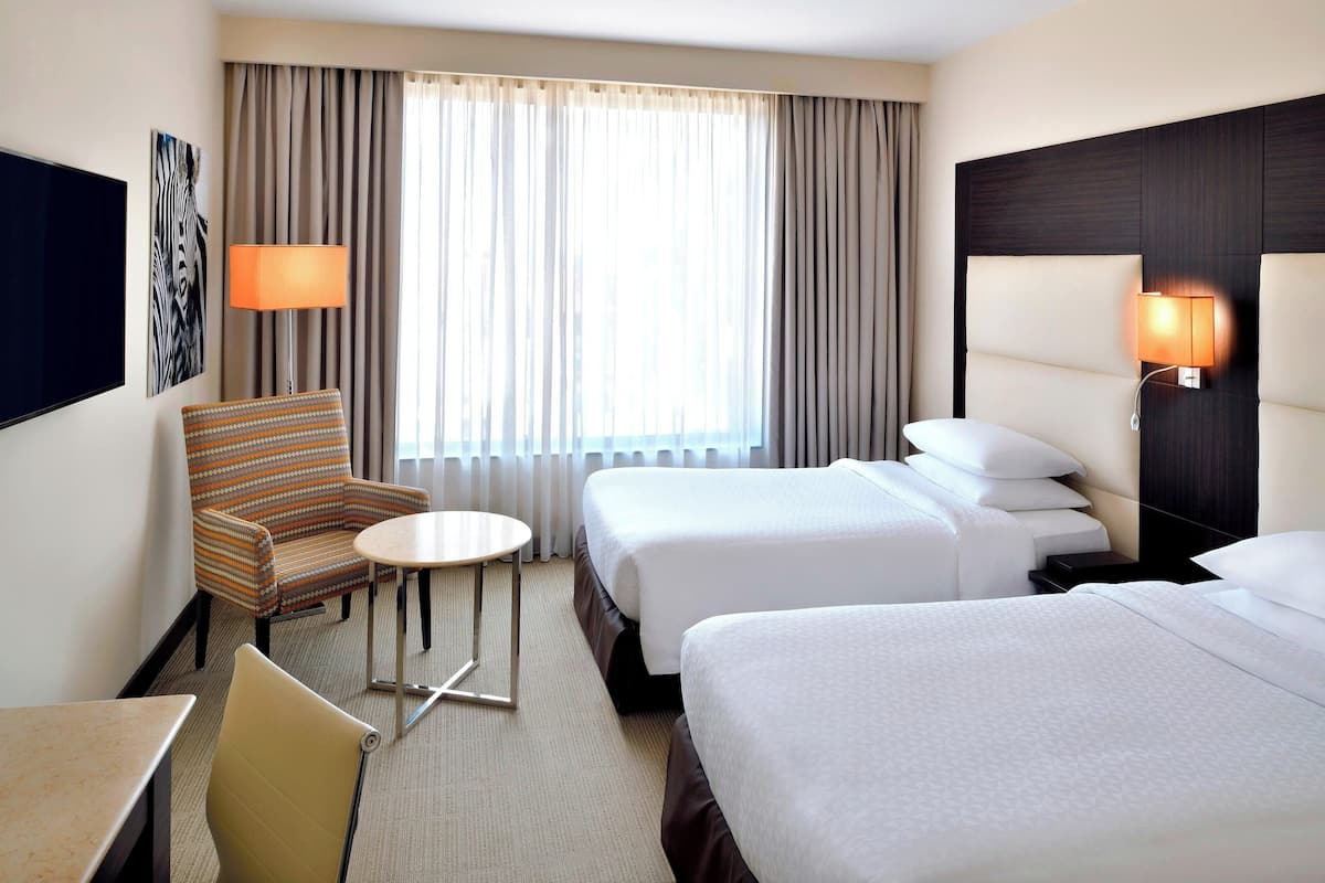 Ein Hotelzimmer mit zwei Betten, einem kleinen runden Tisch, einem Stuhl, einem Fernseher und einer Lampe.