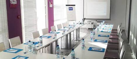 Un salón de conferencias con una larga mesa, sillas, botellas de agua y una pantalla de proyector.