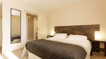 Habitación de hotel con una cama grande, cabecera de madera, dos lámparas de noche, un espejo y una puerta que da a un baño.