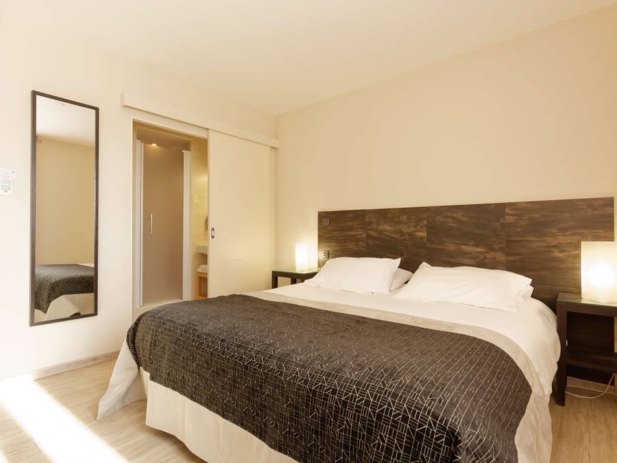 Une chambre d’hôtel avec un grand lit, une tête de lit en bois, deux lampes de chevet, un miroir et une porte donnant sur une salle de bain.
