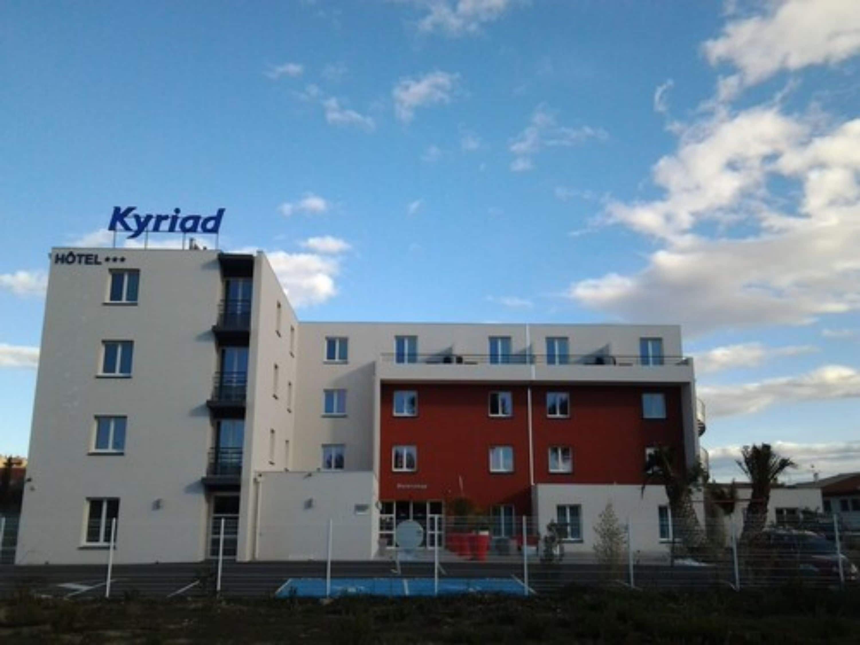 Un hotel llamado 'Kyriad' con un diseño moderno, que presenta una fachada blanca y roja, varios ventanales y un área de piscina visible.