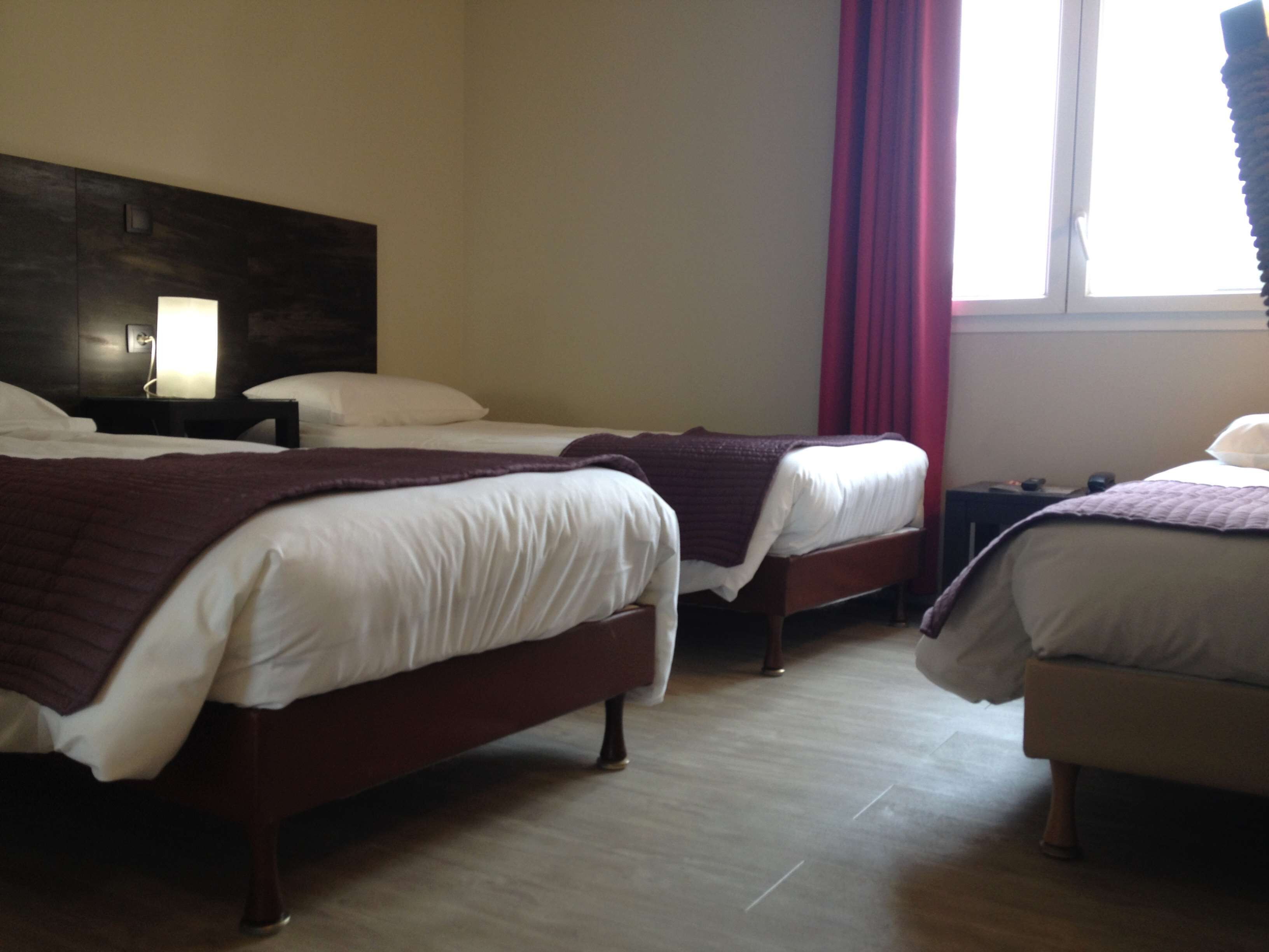 A hotel room with two beds, a wooden headboard, a nightstand with a lamp, and a window with red curtains.