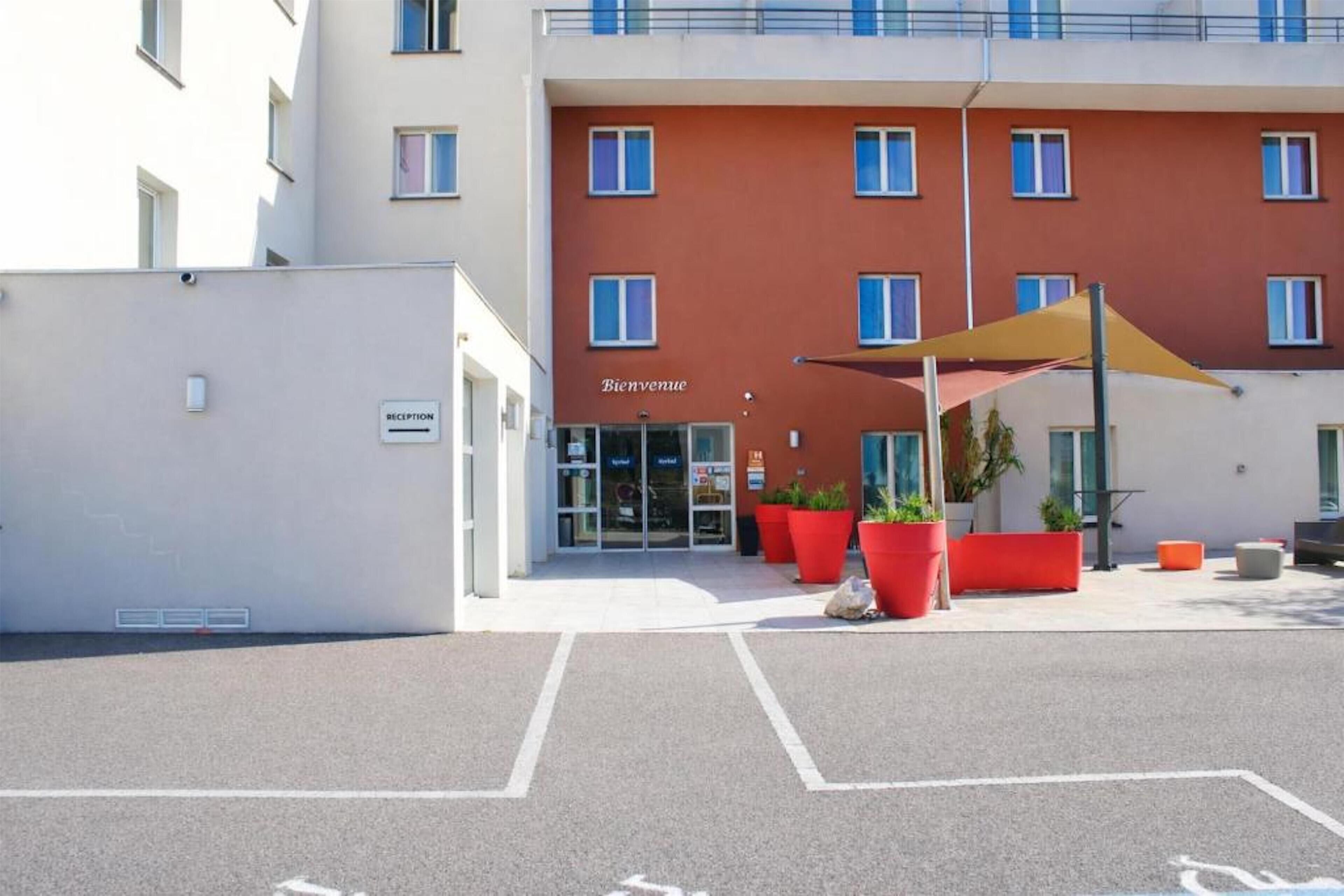 A hotel entrance with a reception desk, a "Bienvenue" sign, and a parking area with marked spaces.
