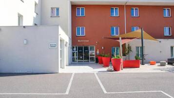 A hotel entrance with a reception desk, a "Bienvenue" sign, and a parking area with marked spaces.