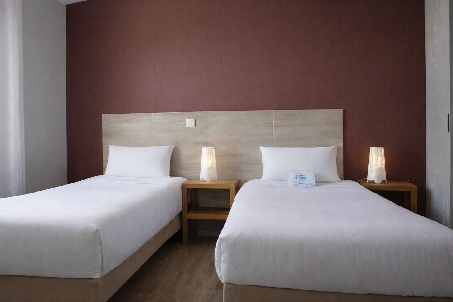 A hotel room with two single beds, wooden headboards, bedside tables with lamps, and a red accent wall.