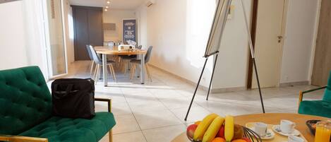 A modern hotel room with a green sofa, a dining table with chairs, a fruit bowl, and a breakfast tray.
