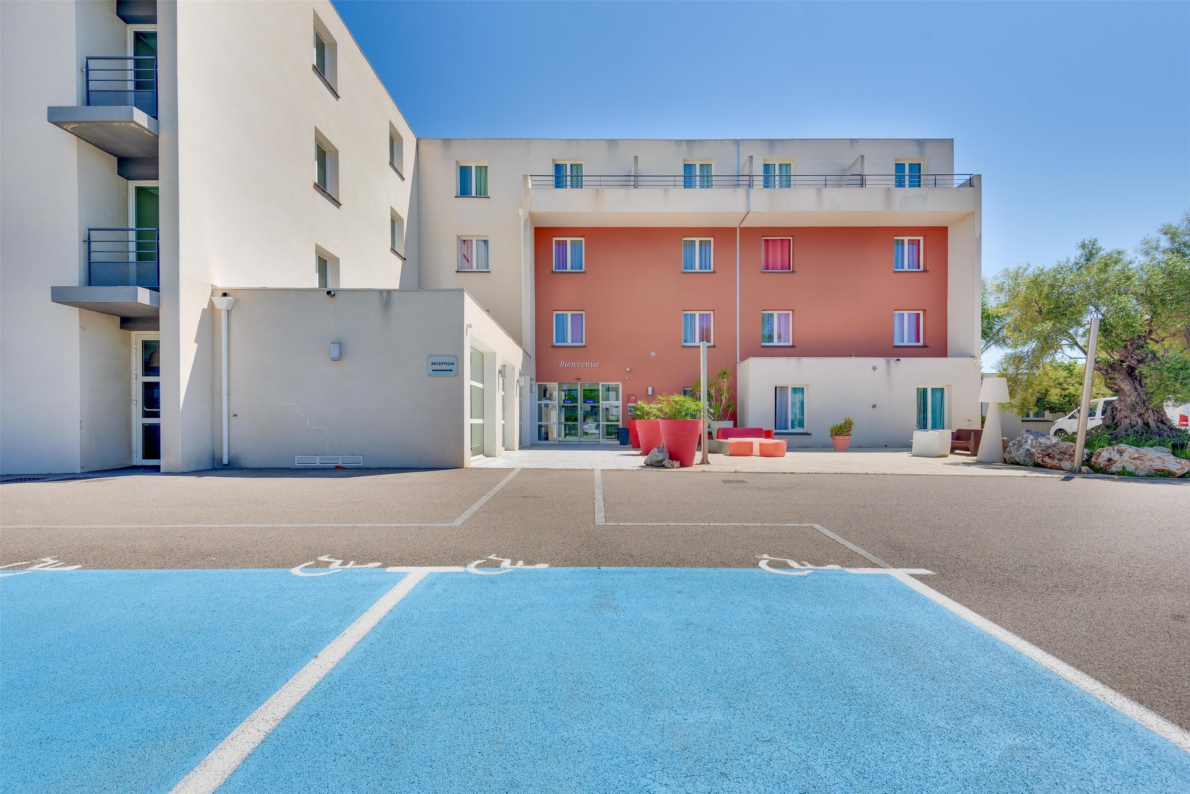 Um edifício moderno com fachada vermelha e branca, área de estacionamento com marcações azuis e plantas em vasos na frente.