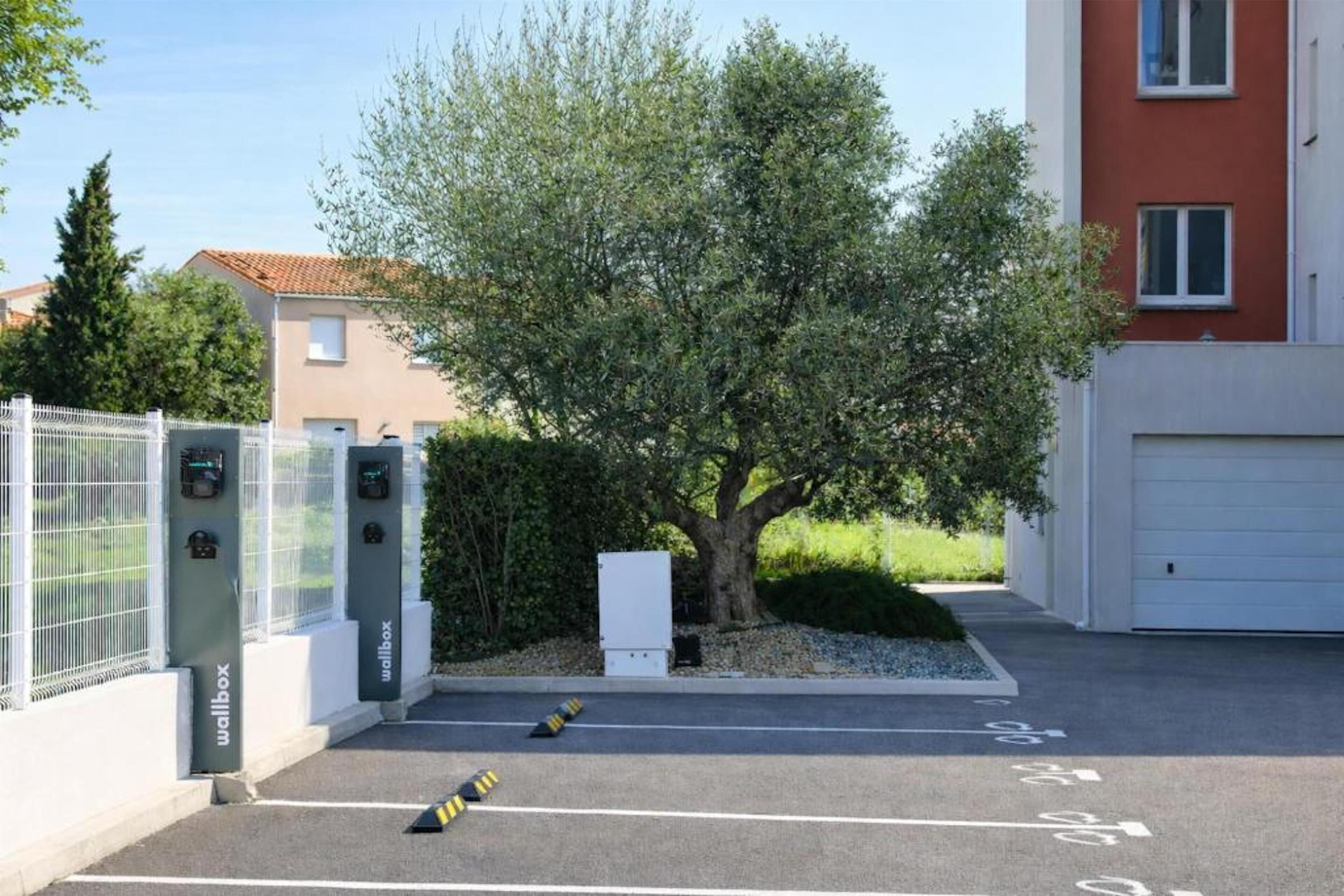 Een parkeerplaats met laadpalen voor elektrische voertuigen, een boom en een gebouw op de achtergrond.