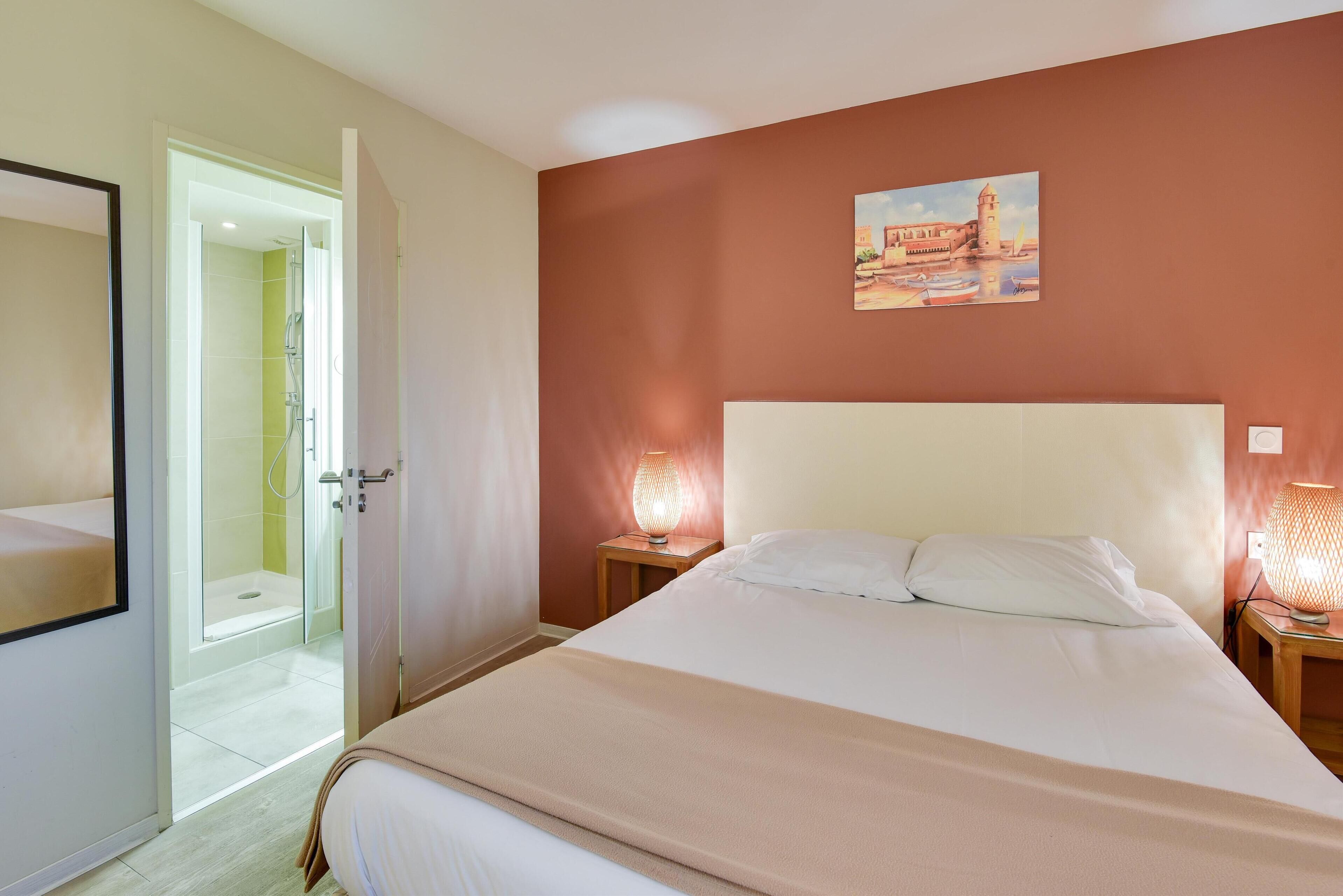 A hotel room with a bed, a bedside table with a lamp, a painting on the wall, and a shower area.