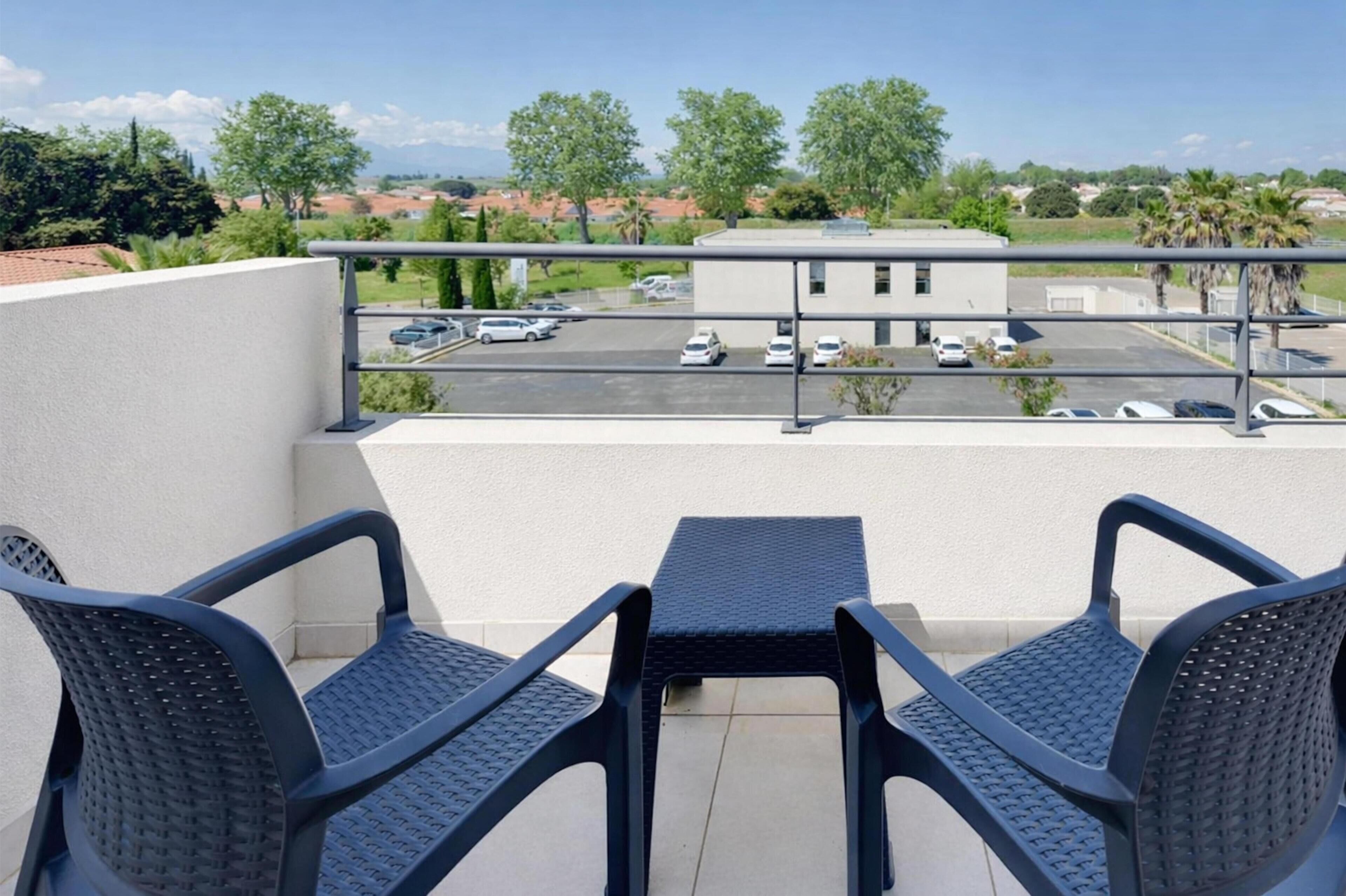 Un balcón con dos sillas y una mesa, con vistas a un aparcamiento y edificios.
