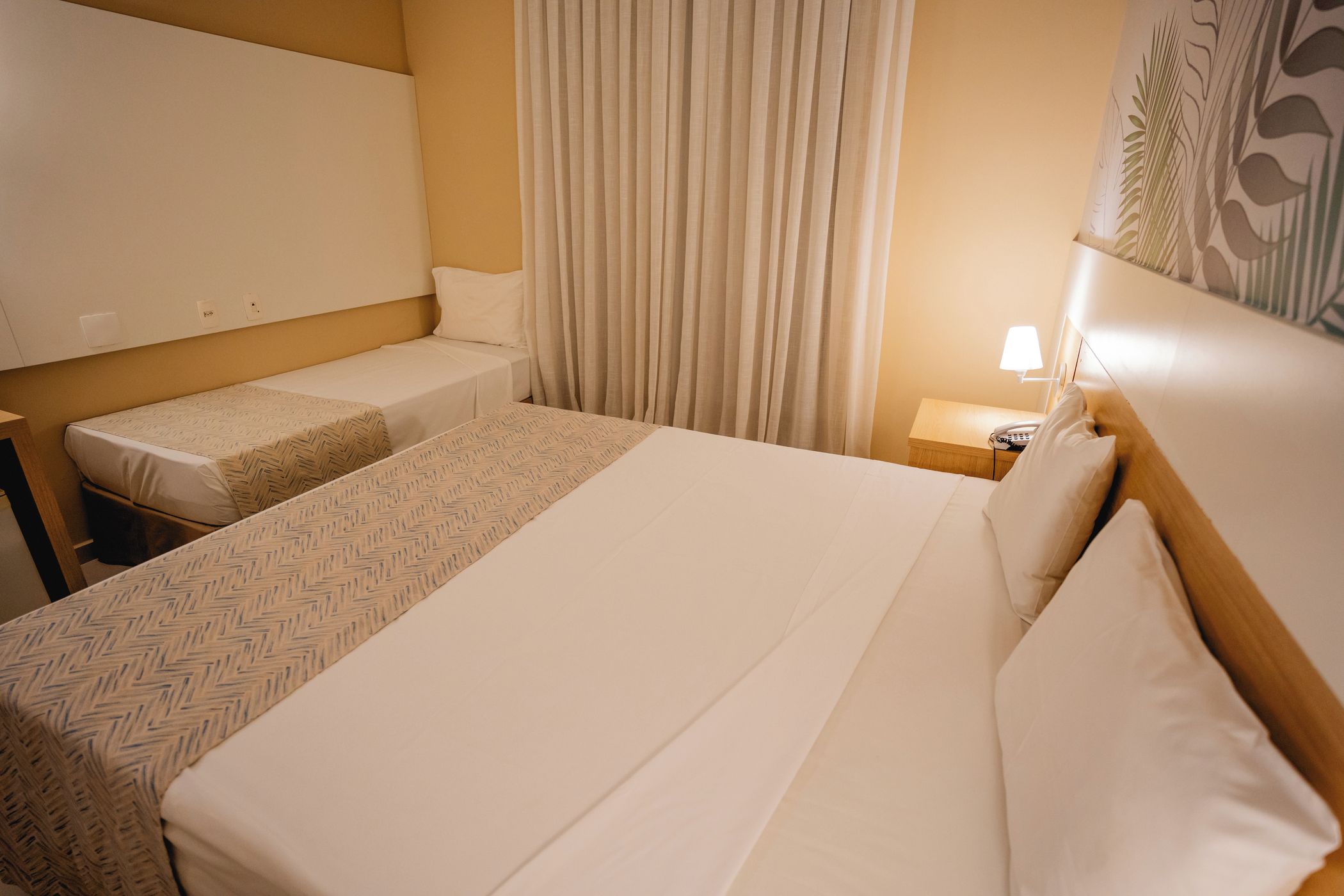 Una habitación de hotel con una cama grande, una cama más pequeña, una mesita de noche con una lámpara y una pared con un diseño de hojas.