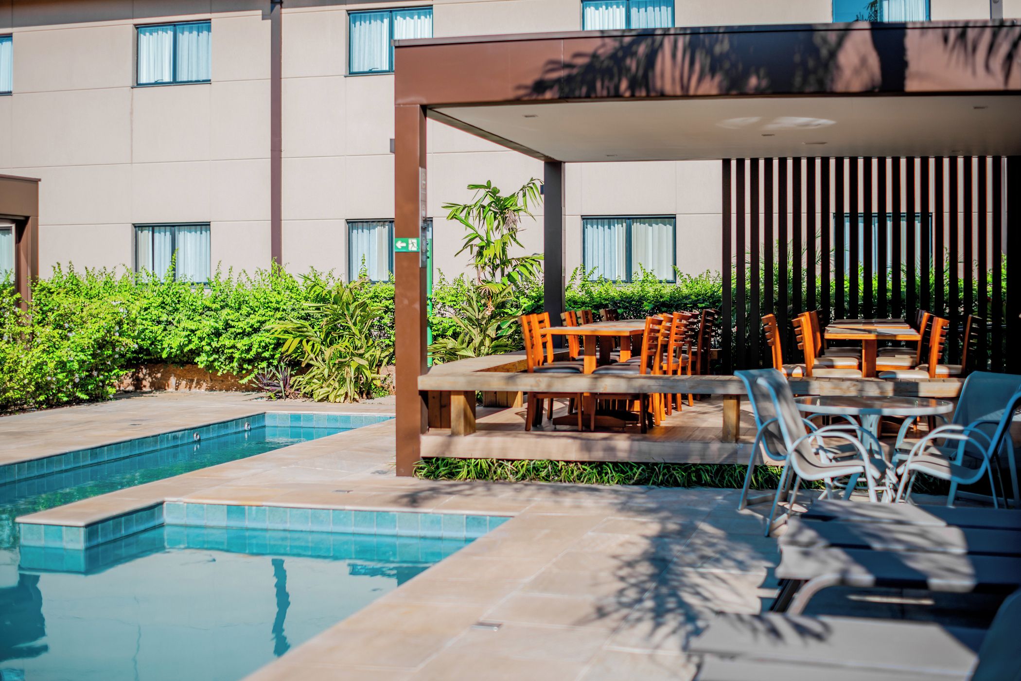 Un área junto a la piscina con una zona de estar cubierta, piscina y un edificio al fondo.