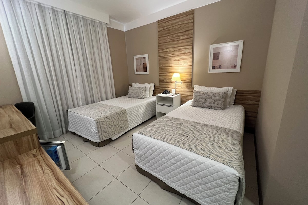 A hotel room with two beds, a desk, a chair, a nightstand with a lamp, and a window with curtains.