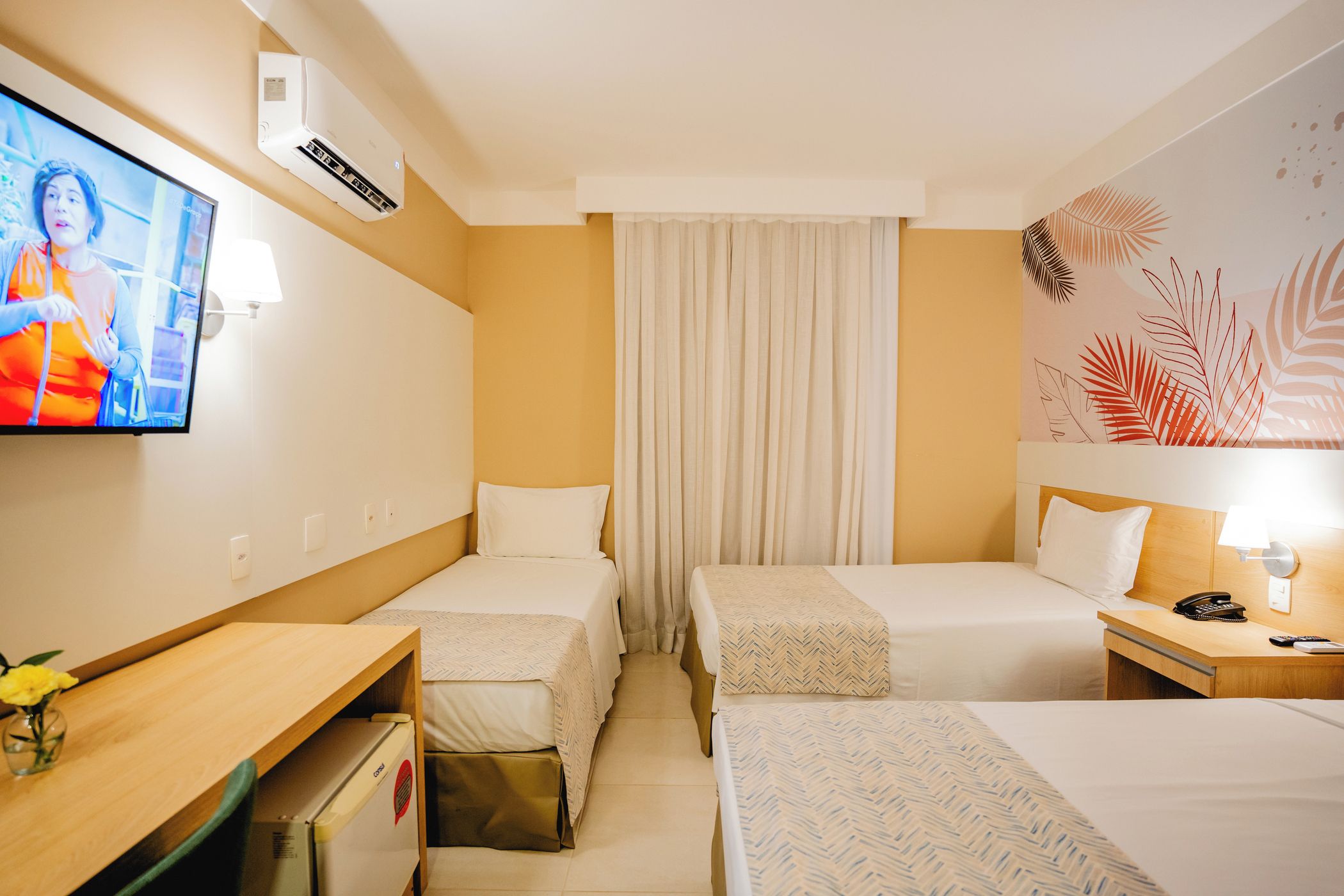 A hotel room with two single beds, a desk with a chair, a television, and a wall-mounted air conditioner.