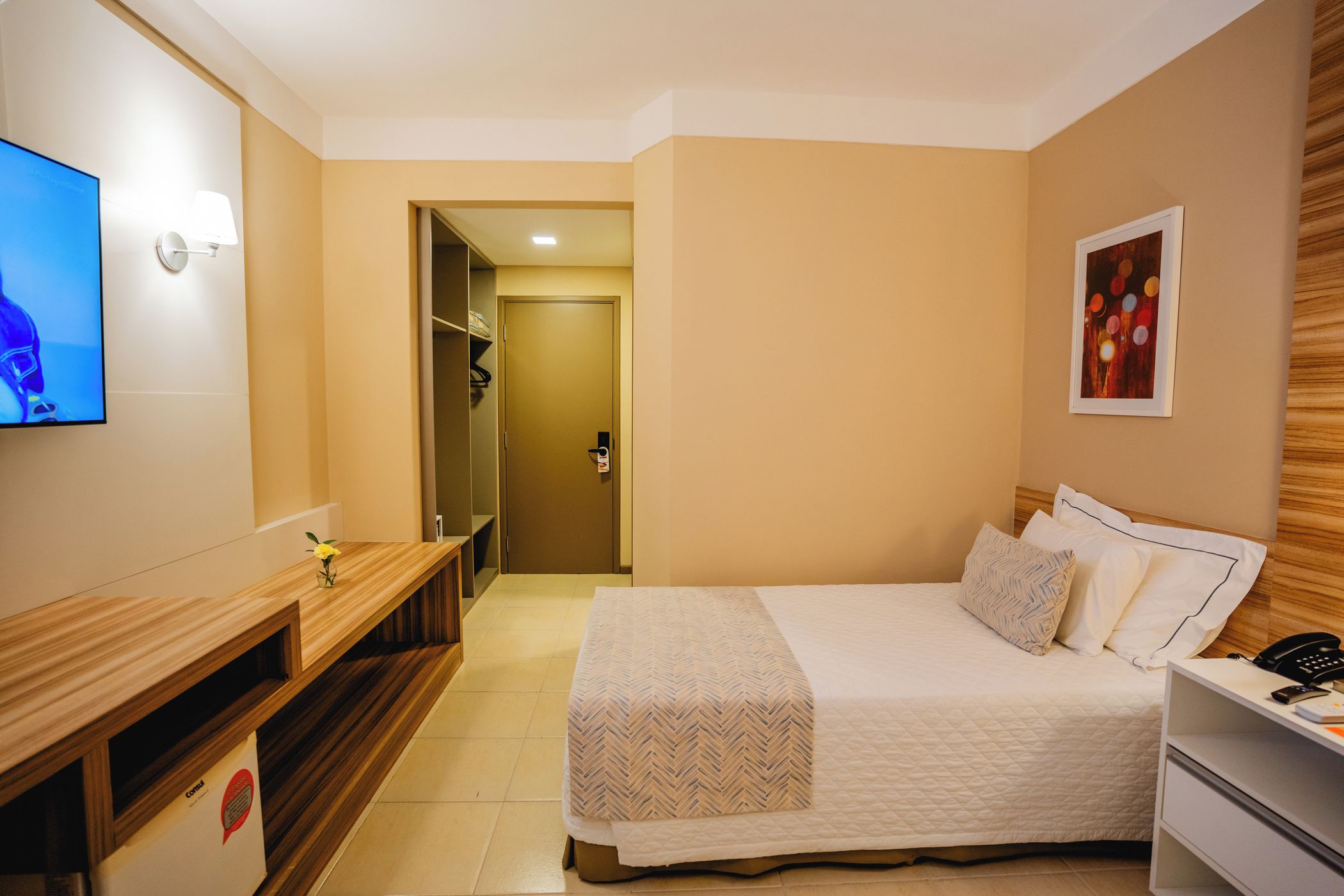 Quarto de hotel com cama, mesa de cabeceira, televisão e escrivaninha com cadeira.