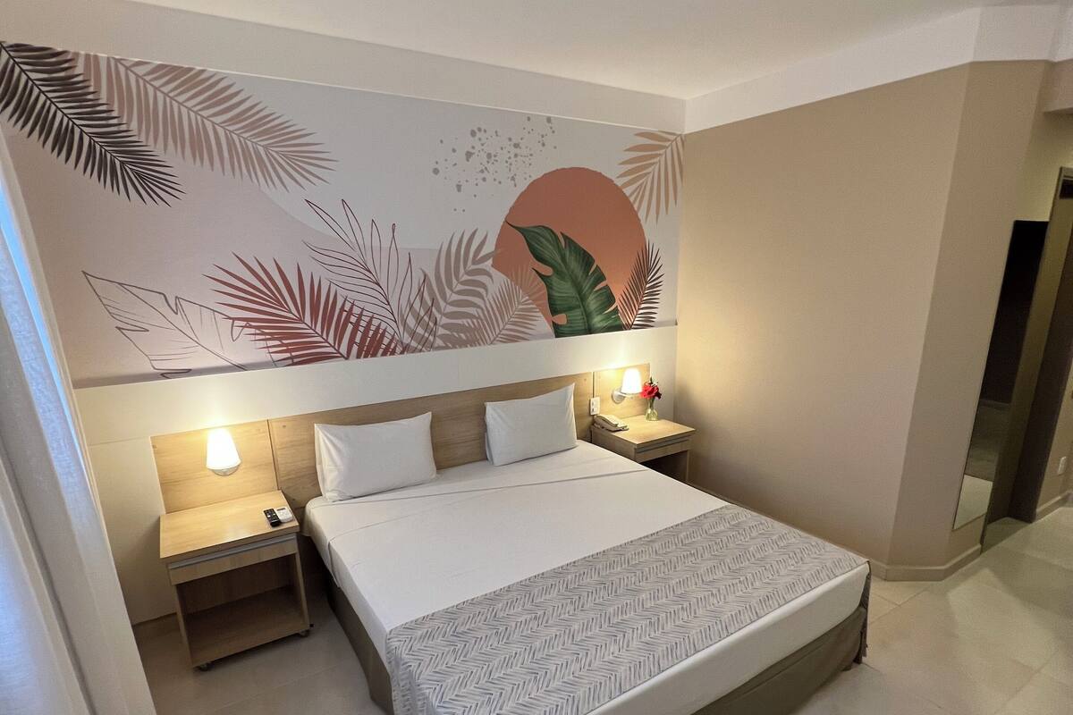 A hotel room with a large bed, a bedside table with a lamp, a small table with a vase, and a chair.