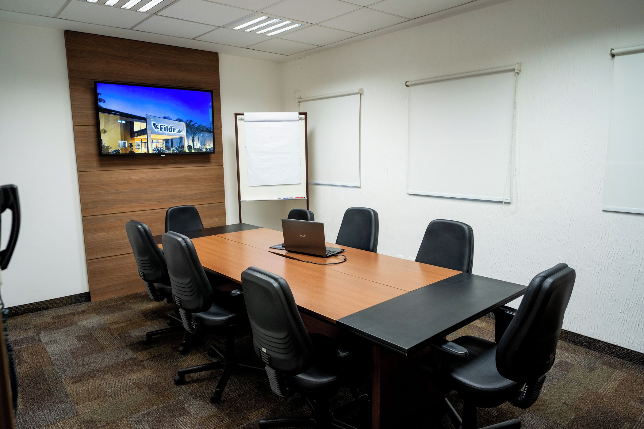 Uma sala de conferências com uma mesa comprida, seis cadeiras, um laptop, uma televisão na parede e dois quadros brancos.