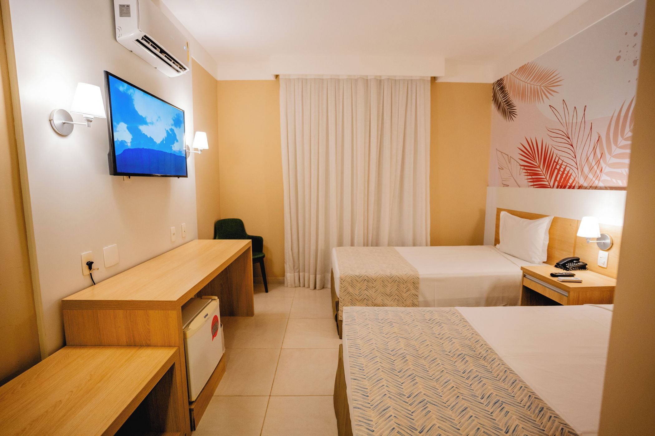 Ein Hotelzimmer mit Schreibtisch, zwei Betten, Fernseher, Telefon und einer Wand-Klimaanlage.