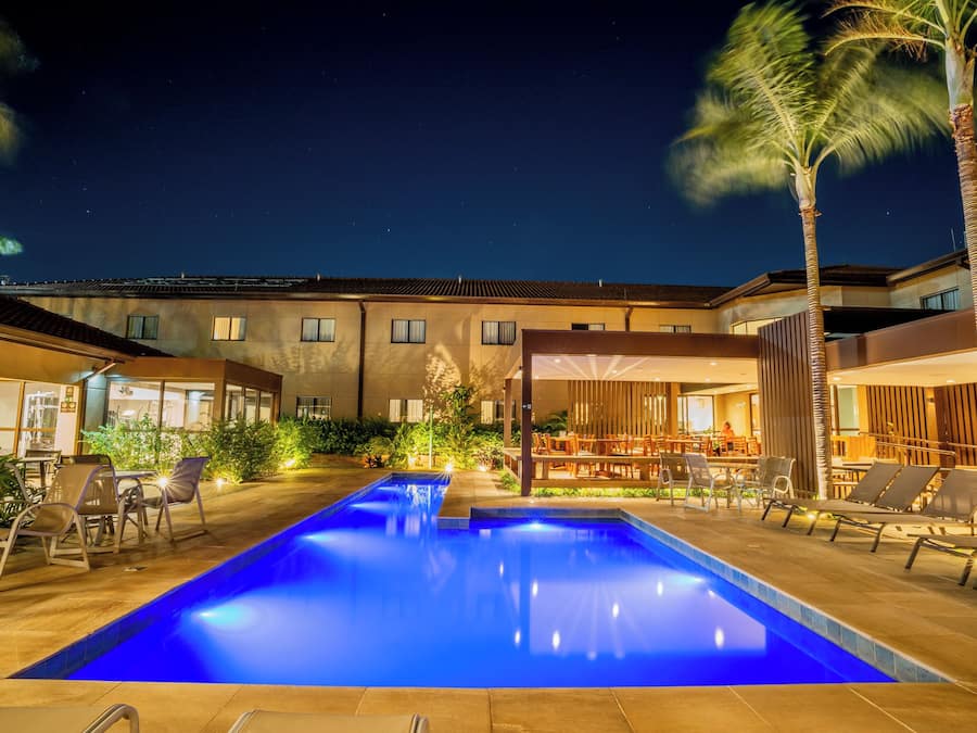 A swimming pool with lounge chairs and tables, surrounded by buildings with large windows and a night sky.