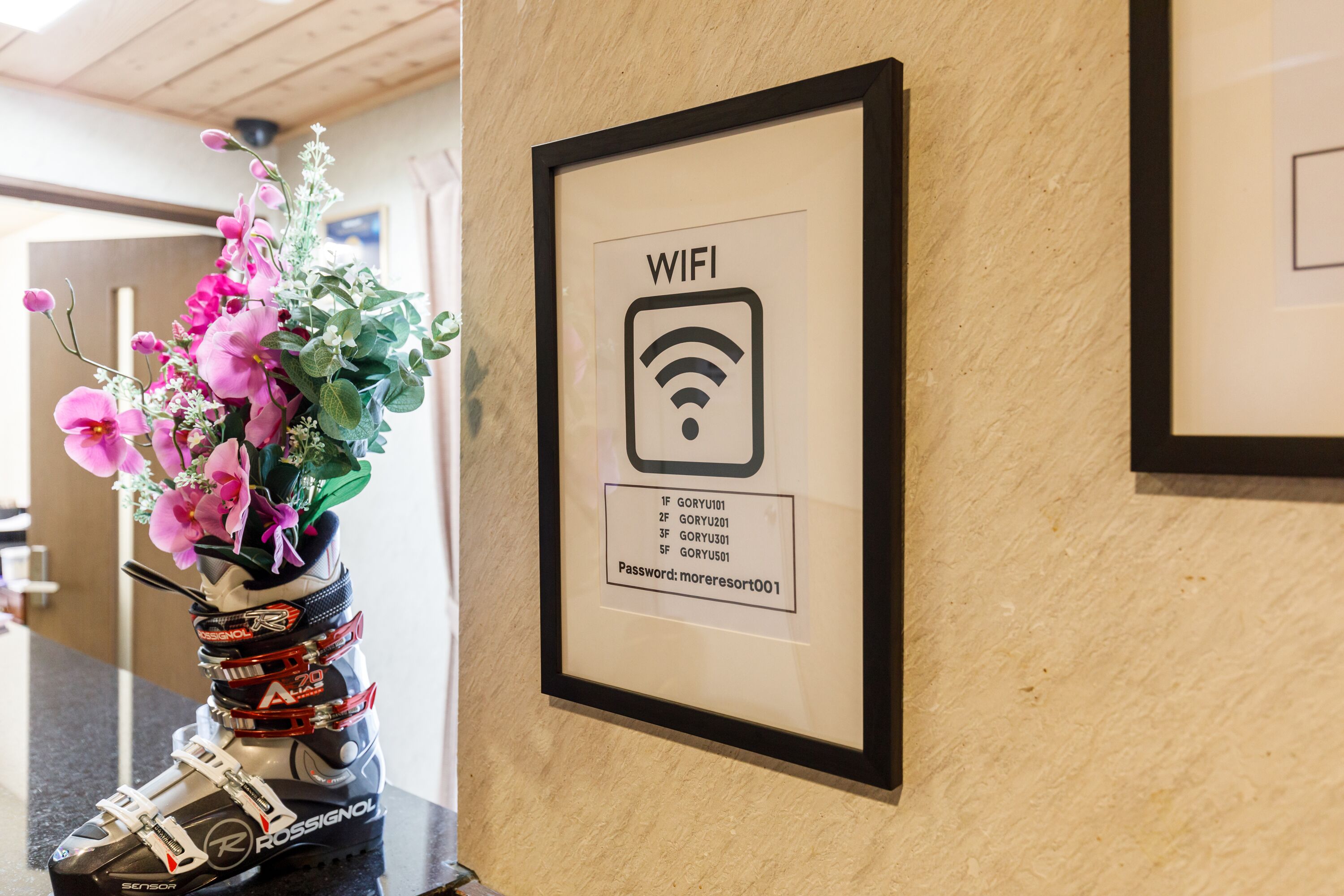 Segnaletica WiFi incorniciata con password su una parete, accanto a un vaso con fiori rosa e attrezzatura da sci.