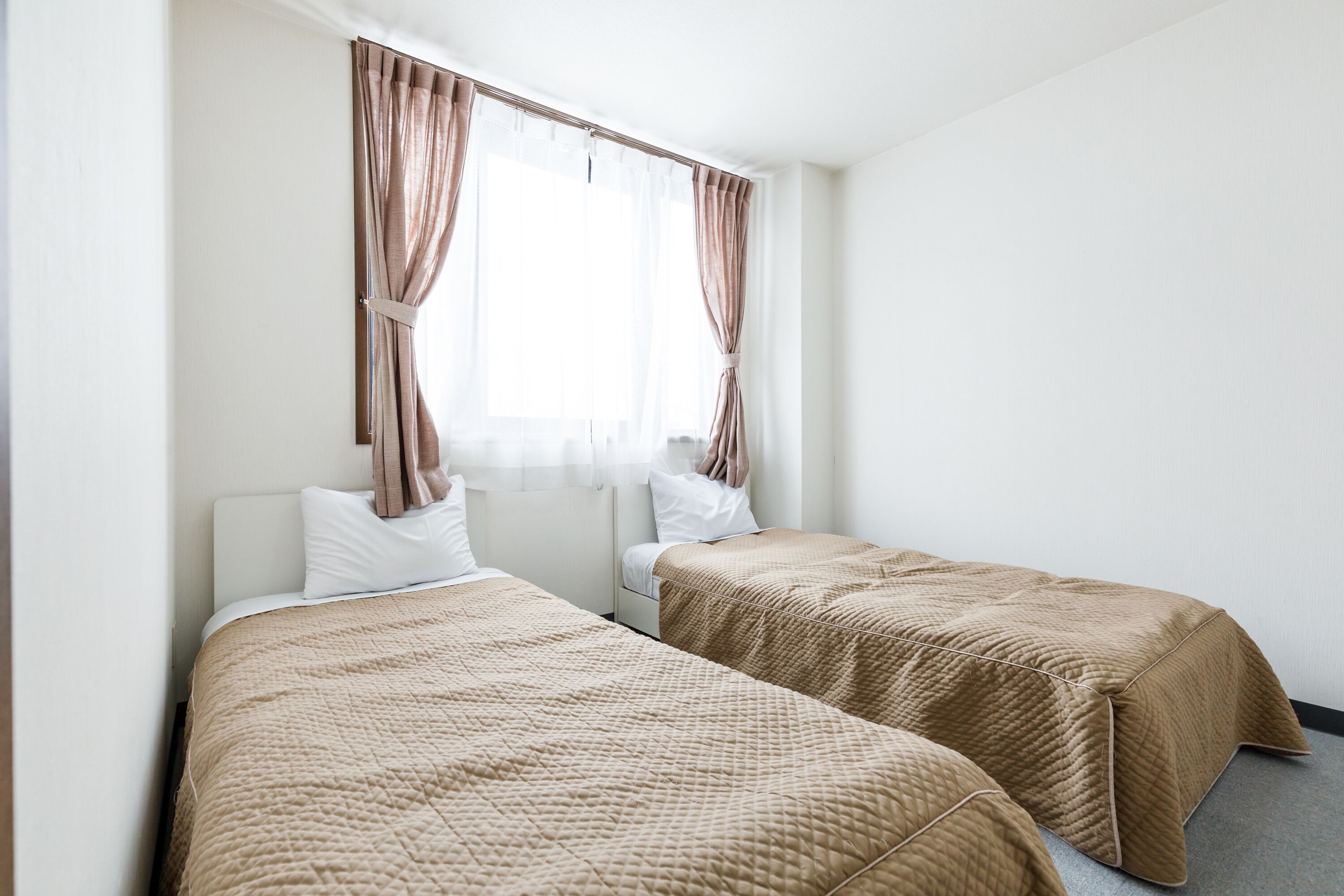 A hotel room with two beds, a window with curtains, and a plain white wall.