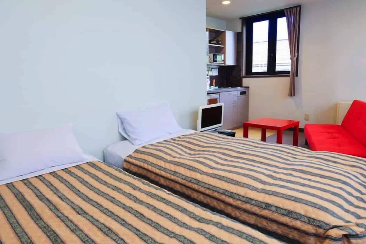 Ein Hotelzimmer mit zwei Betten, einem Schreibtisch, einem roten Sessel und einer Küchenzeile im Hintergrund.
