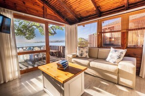 A cozy room with a wooden ceiling, a sofa, a coffee table, and large windows offering a view of the outdoors. - Under Milkwood Resort (Knysna)