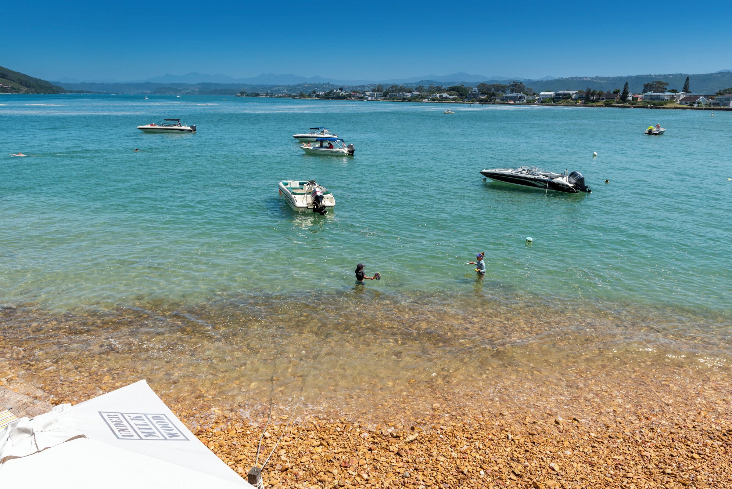 A clear lake with boats, a pebbly shore, and distant hills.