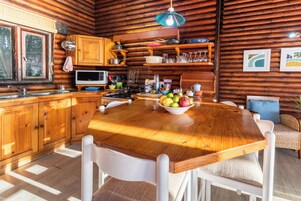 Cocina de cabaña de madera con una mesa de madera, sillas blancas, un cuenco de fruta y varios utensilios de cocina.