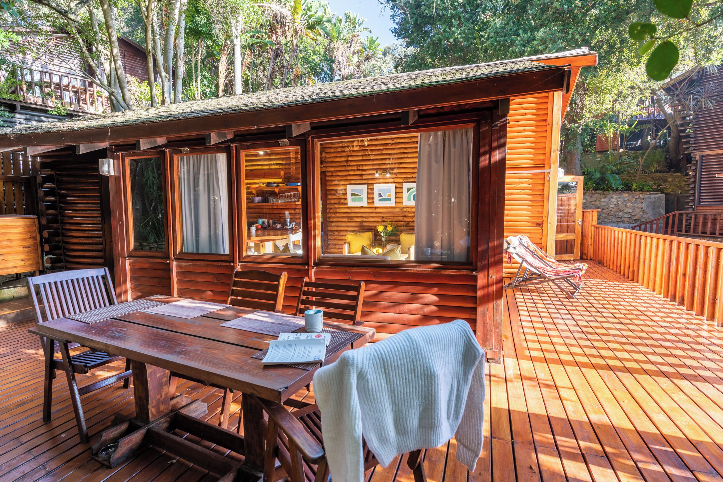 Une cabane en bois comprenant un coin repas, une fenêtre avec des rideaux et une terrasse avec un fauteuil de relaxation.