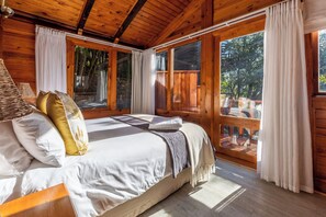 A wooden cabin bedroom with a bed, pillows, and a window overlooking a deck. - Under Milkwood Resort (Knysna)