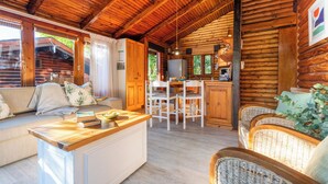 A log cabin interior with a wooden ceiling, a dining area with white chairs, a kitchen with a refrigerator, and a seating area with wicker furniture. - Under Milkwood Resort (Knysna)