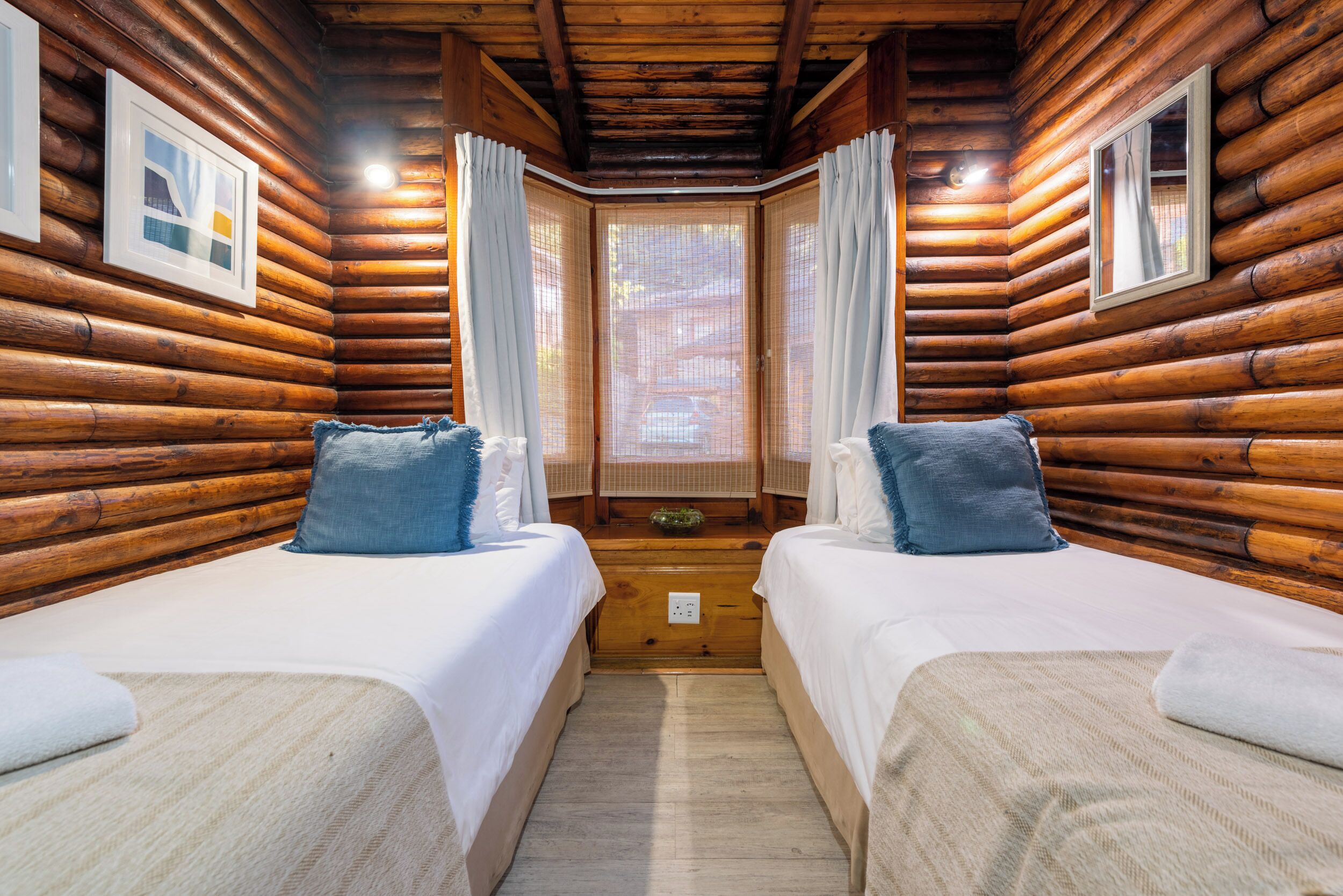 A log cabin room with two beds, a window with curtains, and framed pictures on the walls.