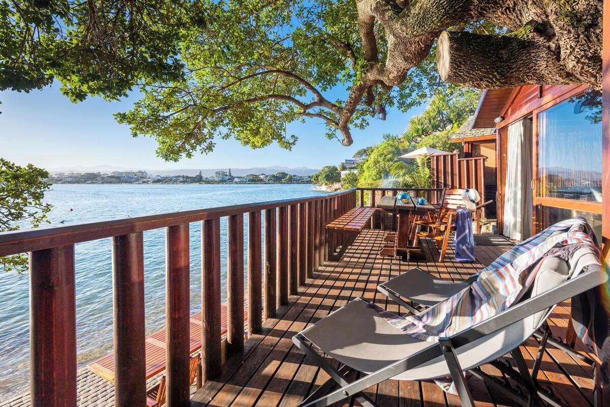 Eine Holzterrasse mit Blick auf das Wasser, ein Tisch mit Stühlen und ein Liegestuhl.