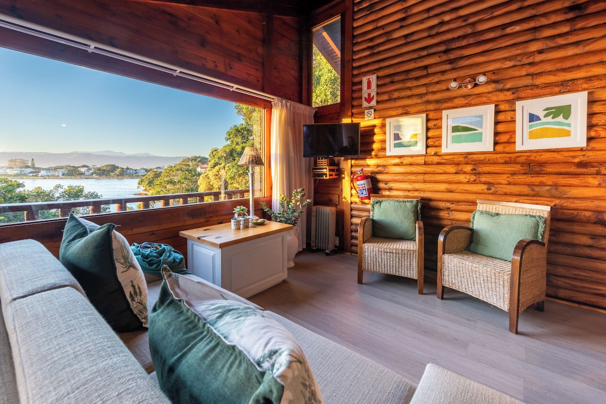 A cozy cabin interior with a wooden exterior, a balcony with a view, wicker furniture, and a mounted television.