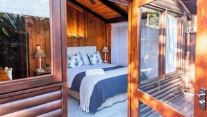 A wooden cabin bedroom with a bed, bedside lamps, and a window. - Under Milkwood Resort (Knysna)