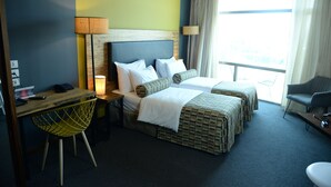 A hotel room with two beds, a desk with a chair, a lamp, and a view of the outdoors. - Royal Court Hotel (Ramallah)