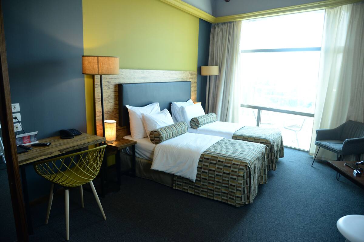 A hotel room with two beds, a desk with a chair, a lamp, and a view of the outdoors.
