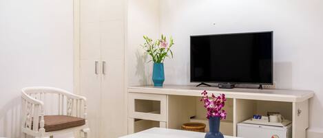 A bedroom with a bed, a white wardrobe, a TV on a stand, a desk with a vase of flowers, and a small refrigerator.