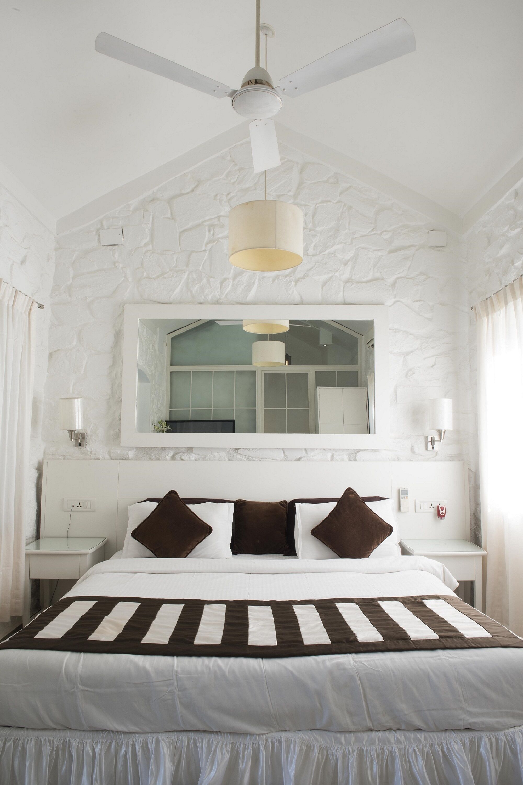 A bedroom with a large bed, a ceiling fan, a mirror, and a stone-textured wall.