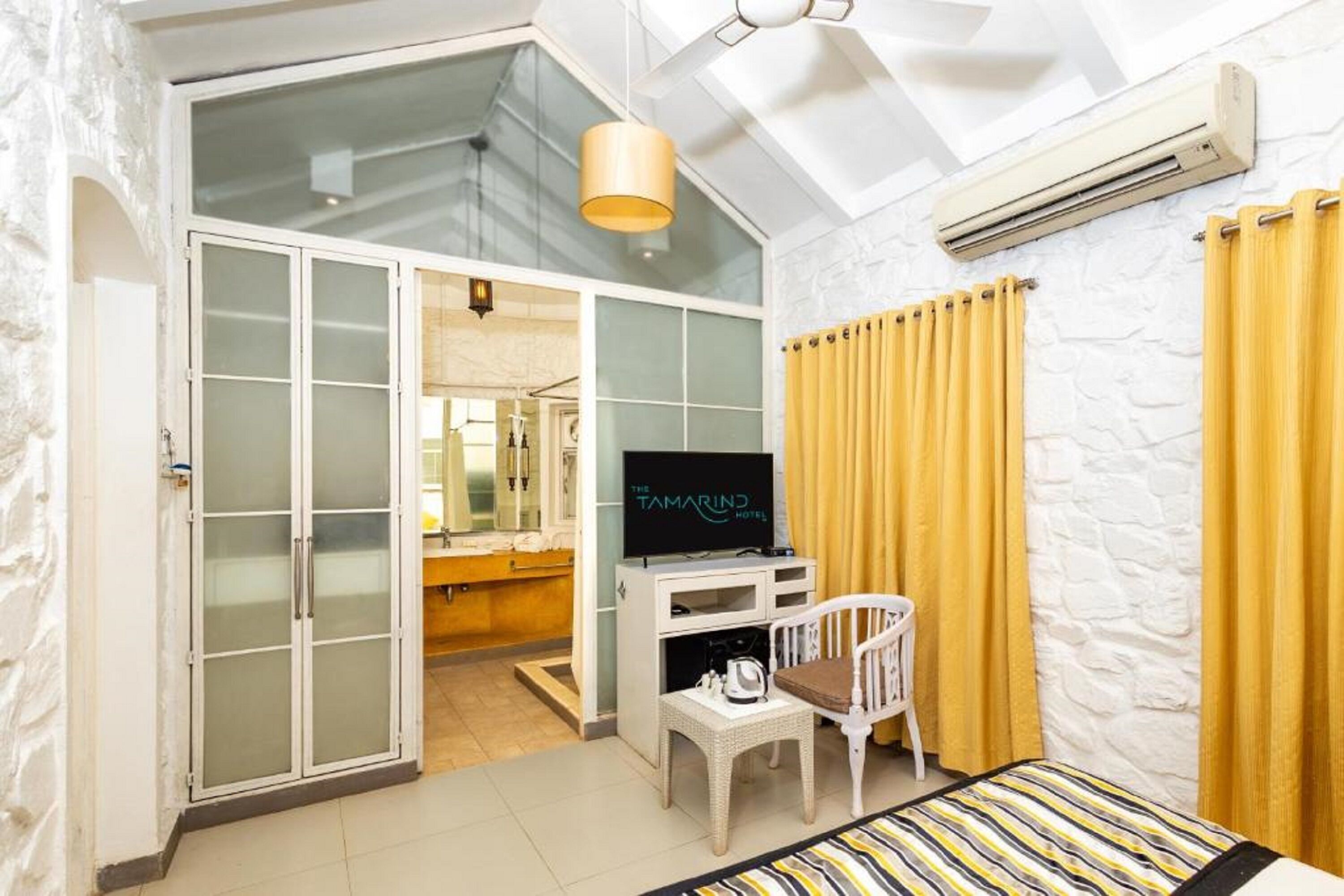 A hotel room with a sliding glass door leading to a bathroom, a flat-screen TV, a small white chair, and a bed with striped bedding.